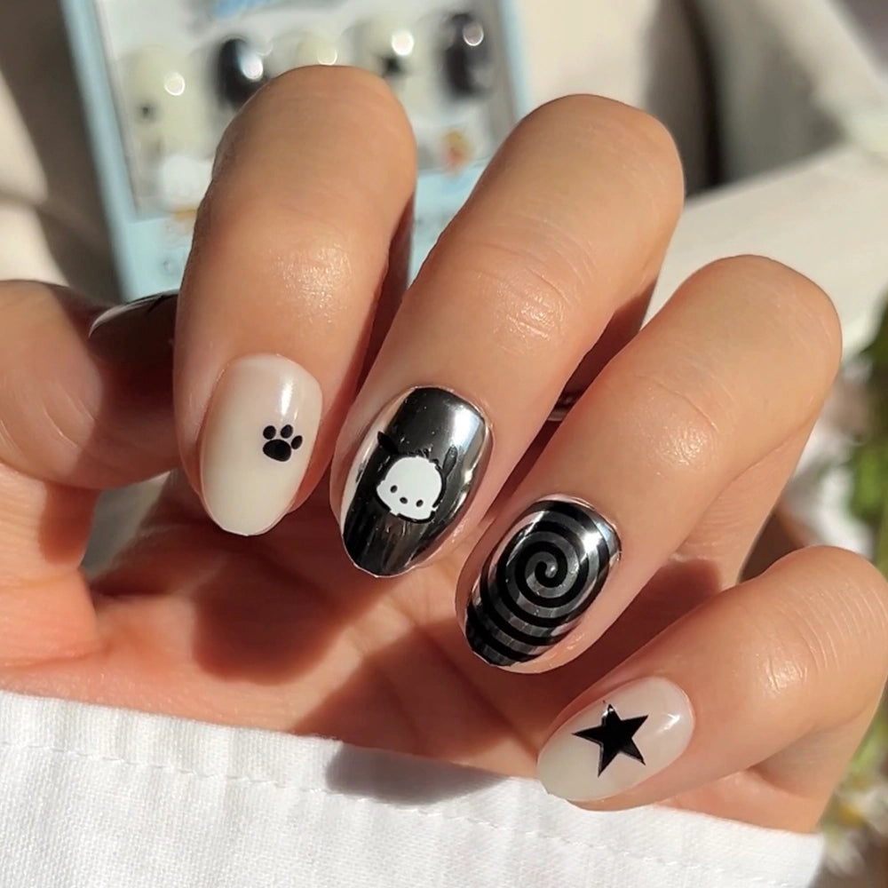 A hand with short oval nails shows off the glamnetic Pochacco™ Nail Set—chrome nails, a black paw print, a cartoon dog face, spiral, and star on nude and metallic backgrounds that shine in natural light.