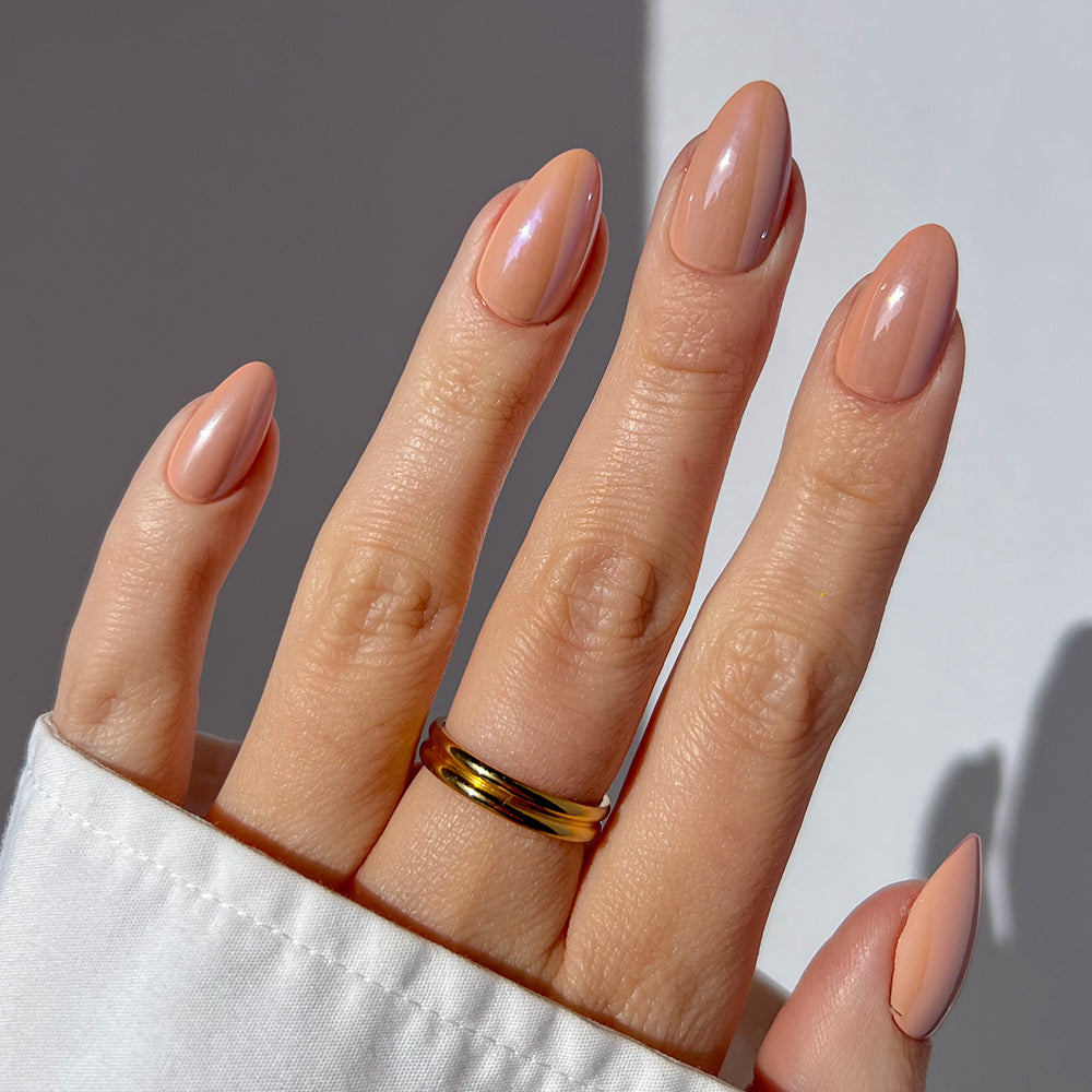 A hand with short almond nails from glamnetic’s "Los Angeles" nude-blush set shows off a shiny gold ring on the ring finger, posed against a light background and styled with a white long-sleeve shirt. - Product image