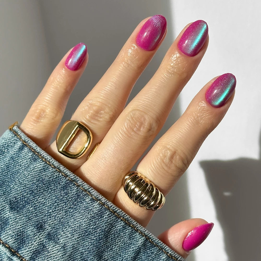 A hand with shiny pink and blue iridescent short oval nails wears two chunky gold rings—one D-shaped, one ridged. The velvet metallic finish of glamnetic’s Berry Fizz nails stands out against a blue denim jacket.