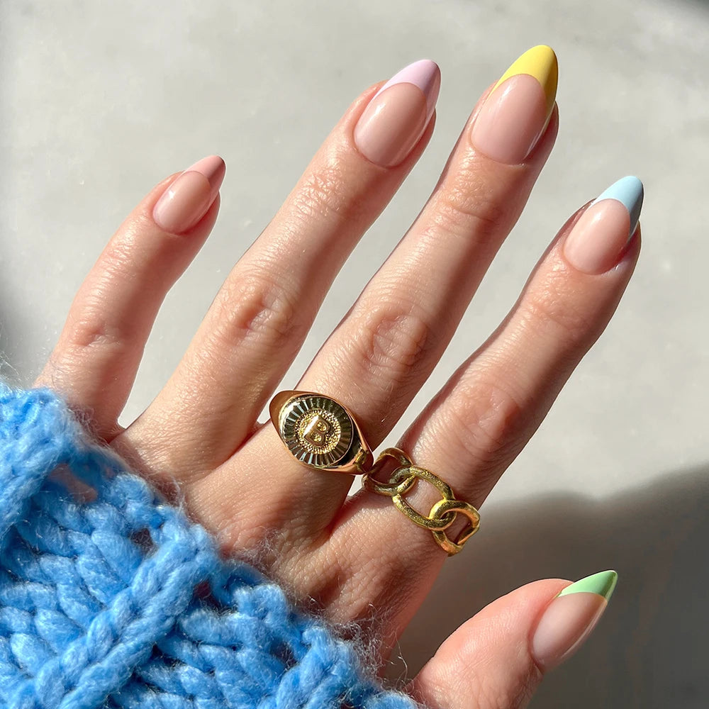 A hand with short almond nails in glamnetic's Confetti pastel French tips is held up to a light background, accessorized with a chunky blue knit sleeve, a gold B emblem ring, and a gold chain ring.