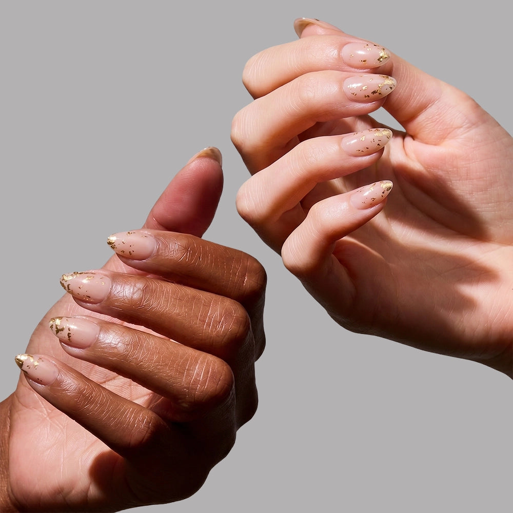 Two hands with manicured short almond nails, featuring a natural nude base and gold foil accents from glamnetic's 24K set, are shown against a plain gray background. The hands have different skin tones and are posed gracefully. - Product image