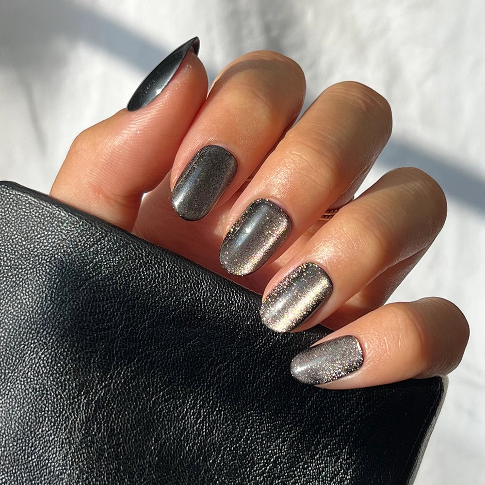 A hand with short round nails painted in glamnetic's Galactic, a shiny metallic dark gray polish with subtle glitter, holds a textured black leather item.