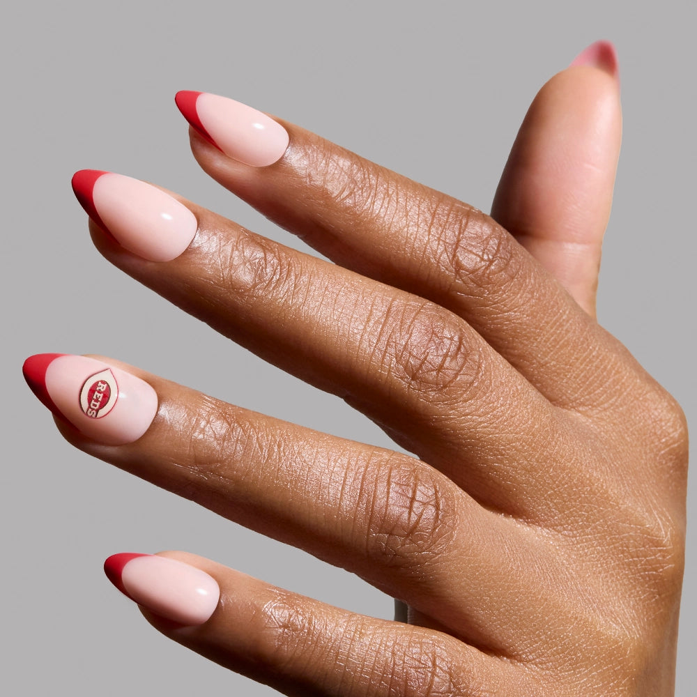 A hand showcases glamnetic's Cincinnati Reds Press-On Nail Set with almond-shaped, nude nails, bold red French tips, and a thumb featuring a red circle with "RED" inside, all against a plain gray background.