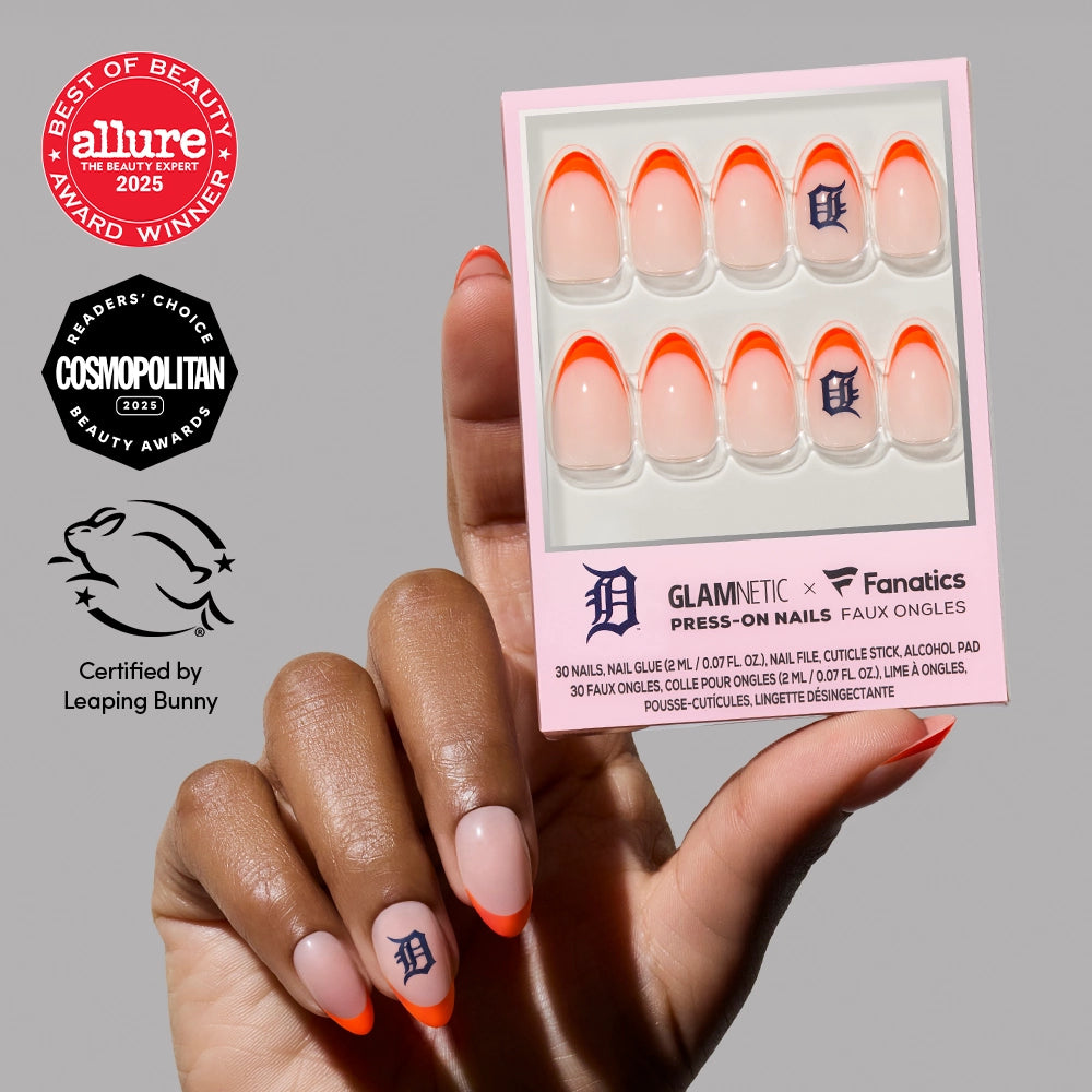 A hand with long, almond-shaped nails—orange French tips and blue "D" accents—holds a Detroit Tigers Press-On Nail Set by glamnetic. Award and certification logos appear on the left.