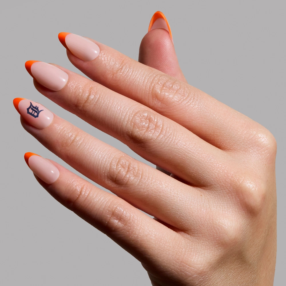 A hand displays almond-shaped nails with a nude base, bright orange French tips, and a black symbol—perfect for Detroit Tigers fans. Try the glamnetic Detroit Tigers Press-On Nail Set, shown here on a plain gray background.