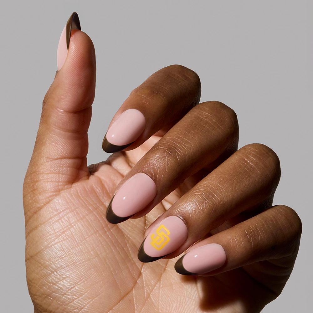 A hand wears glamnetic's San Diego Padres Press-On Nail Set, featuring almond nails in light pink with dark brown tips and a yellow geometric accent. A subtle fan-inspired look is shown against a plain gray background.