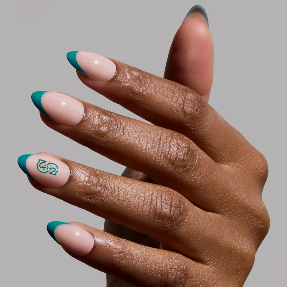 A hand with almond-shaped Glamnetic Seattle Mariners Press-On Nails features a nude base, teal French tips, and a ring finger with a teal abstract design. The plain gray background highlights the Glamnetic nail set.
