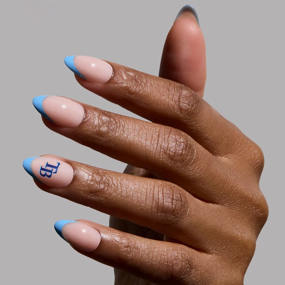 A hand displays the glamnetic Tampa Bay Rays Press-On Nail Set: short almond press-ons with nude and light blue French tips, plus blue "TB" letters on the ring fingernail, shown against a plain gray background.