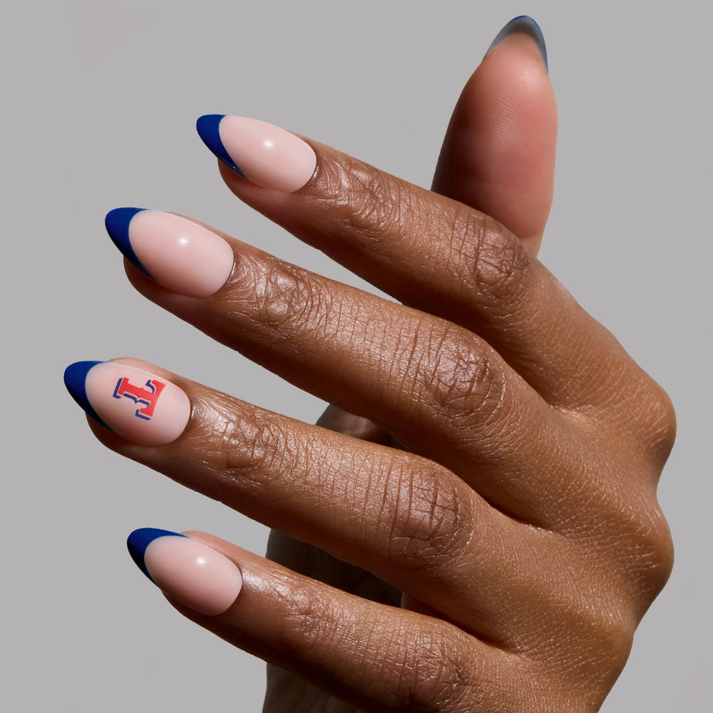 A hand with short almond nails has a nude base, bold blue French tips, and a red and blue letter J on the ring fingernail. Featuring the glamnetic Texas Rangers Press-On Nail Set against a plain grey background.