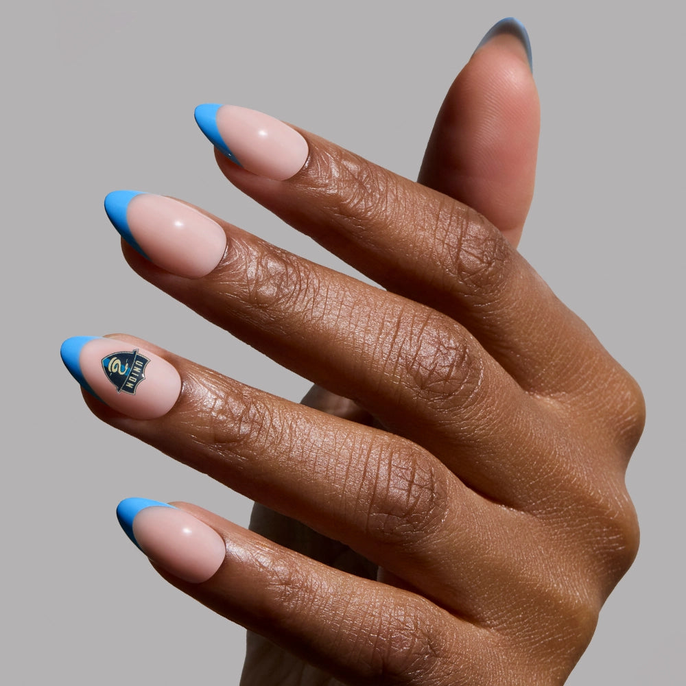 A hand with short almond nails wears the glamnetic Philadelphia Union Press-On Nail Set, showing a nude base and bright blue French tips; the ring fingernail has a small black and green geometric design. The background is solid light gray.