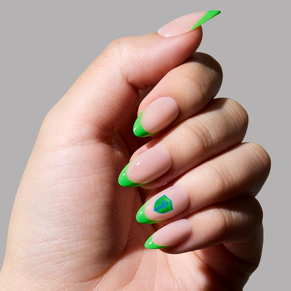 A hand with almond-shaped nails featuring neon green tips and a geometric accent on the ring finger, styled as the Seattle Sounders Press-On Nail Set by glamnetic, shown against a plain light background.