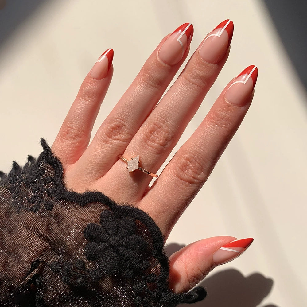 A hand with short almond nails wears glamnetic’s Red Martini press-ons, showcasing a festive red and white geometric French manicure. A lace sleeve and a gold ring with a clear gemstone complete this elegant, holiday-ready look.