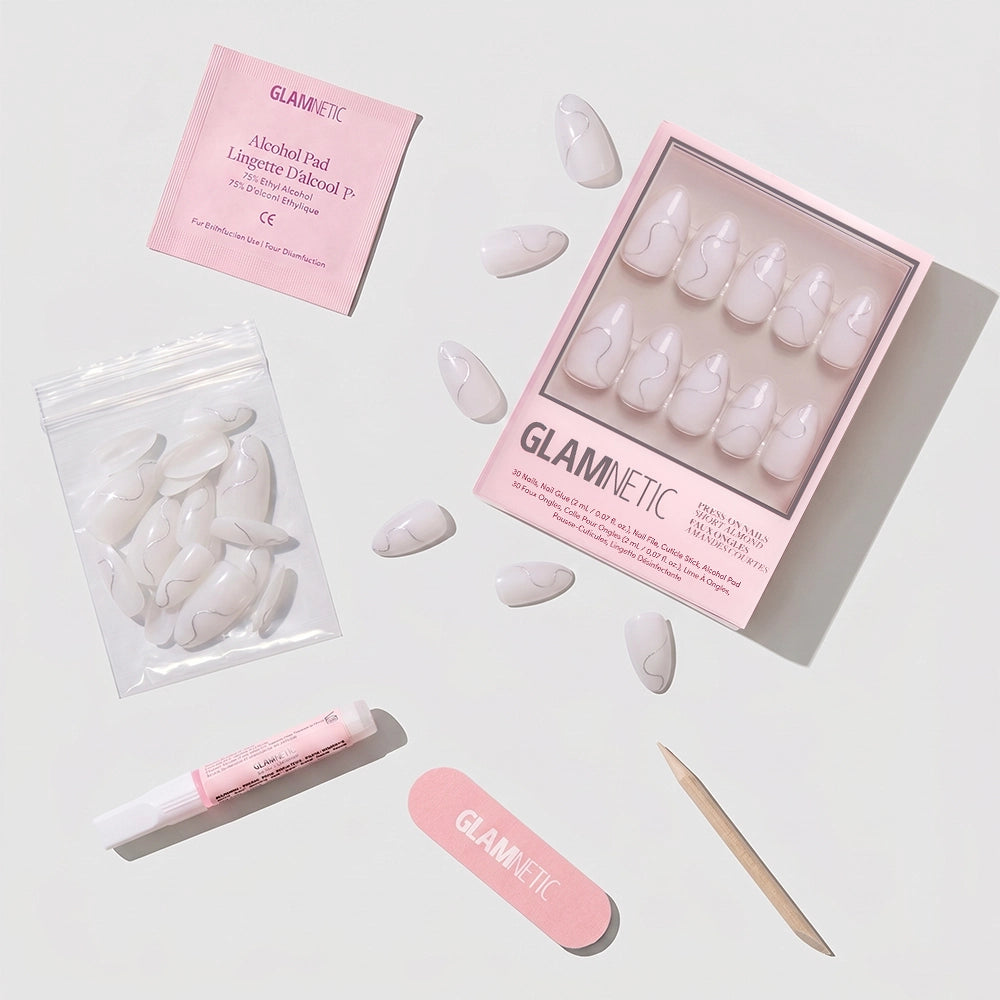 Flat lay of the glamnetic Silent Night press-on nails kit, featuring short almond white nails, a pink nail file, wooden cuticle stick, nail glue, an alcohol pad, and extra vegan nails in a plastic bag on a white background. - Product image
