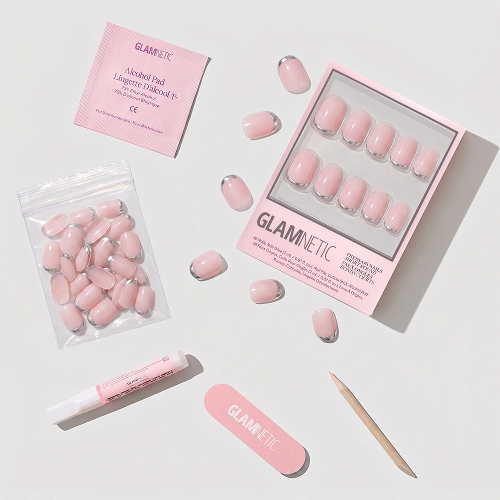 A flat lay of glamnetic's Silver Belle press-on nails, pink nail file, wooden cuticle stick, glue tube, alcohol pad, and extra short round nails in a clear bag, all neatly arranged on a white background. - Product image