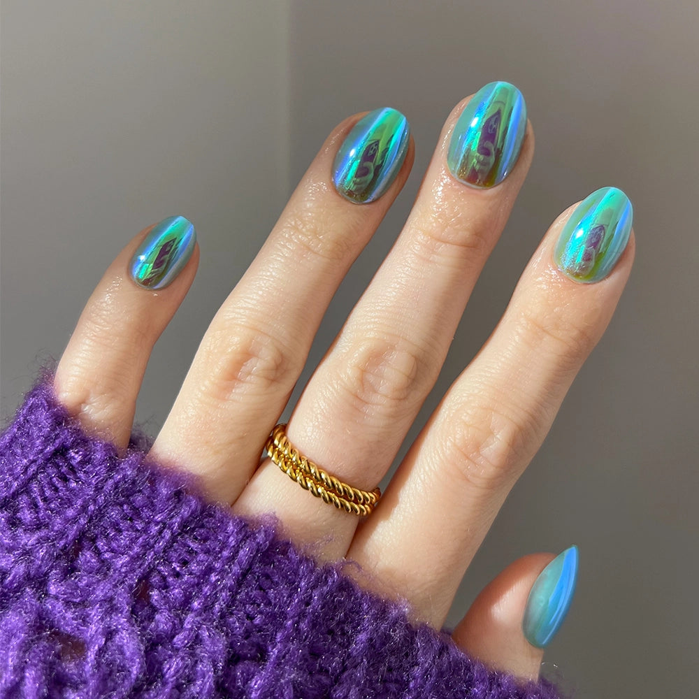 A hand with long, almond-shaped nails from glamnetic's Siren set shows a shiny, iridescent blue-green chrome finish. A gold chain ring adorns one finger, and a textured purple sweater sleeve partially covers the hand.