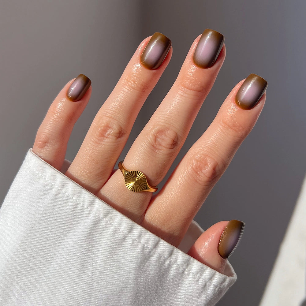 A hand wears glamnetic’s Eclipse taupe brown nail set and pink ombré squoval nails, paired with a gold fan-shaped ring and a white long-sleeve shirt.