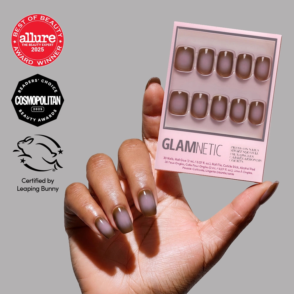 A hand with glossy, short squoval Eclipse press-on nails by glamnetic holds a package of matching taupe brown fake nails against a gray background, showing beauty award logos and Leaping Bunny cruelty-free certification.