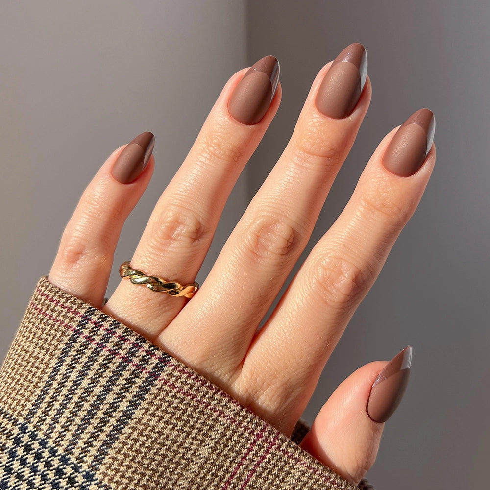 A hand with glamnetic's Terra vegan matte almond nails in brown, wearing a gold twisted ring, extends from a plaid sleeve in beige, brown, and dark red tones.