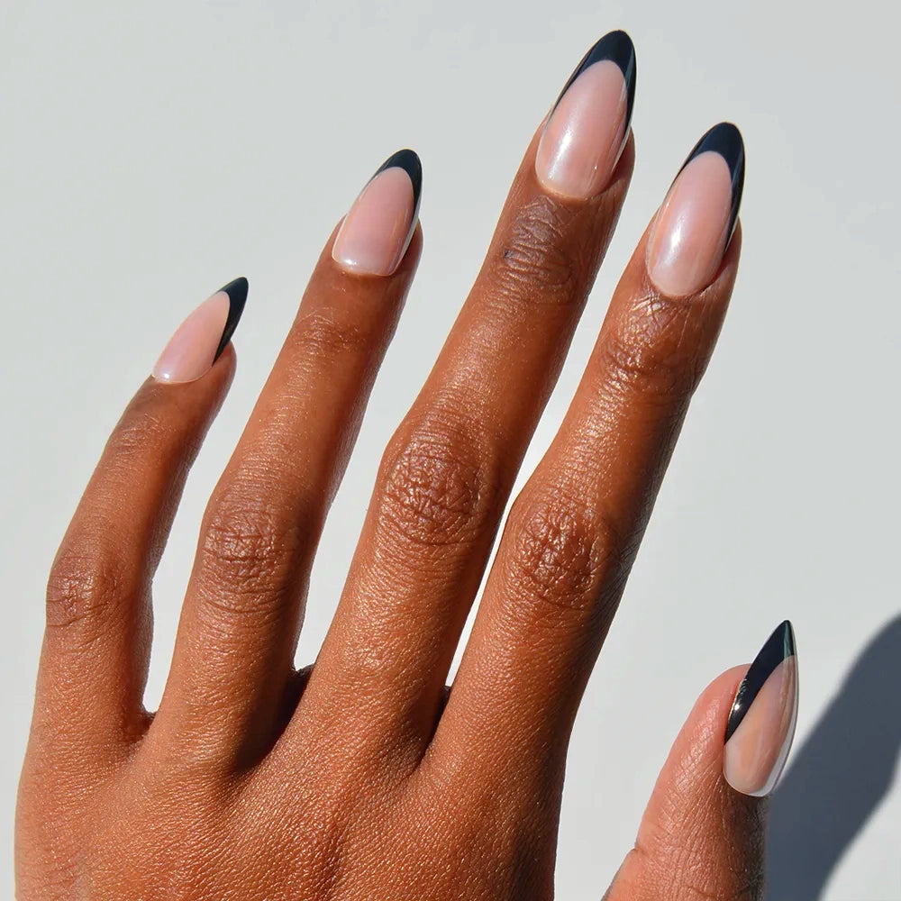 A hand with almond-shaped nails wears glamnetic's Black Truffle press-ons, showcasing a minimalist black French tip on a natural glossy base, set against a plain light background.
