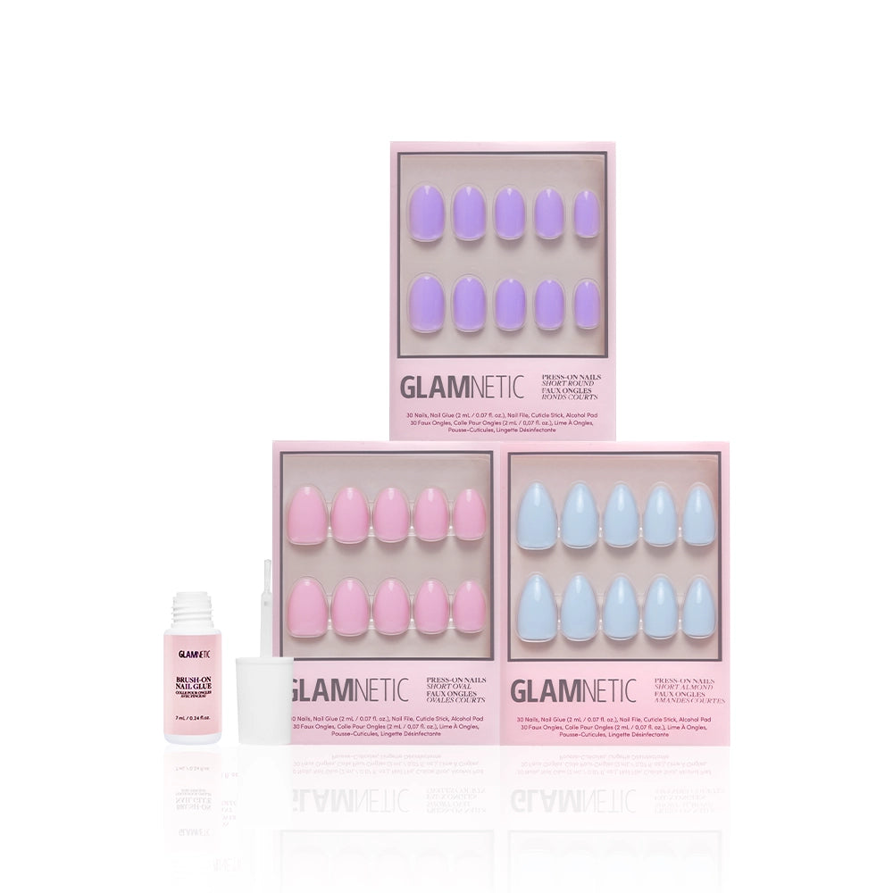 Three boxes of glamnetic Blooming Buds press-on nails in pastel purple, pink, and blue short almond shapes are stacked in a pyramid, with a small white bottle of nail glue and its open cap placed in front on a white background. - Product image