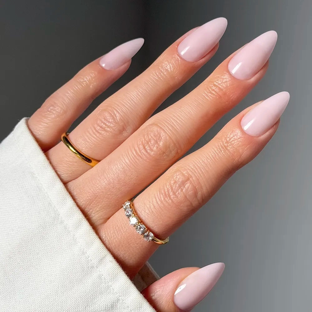 A hand with long, almond-shaped glamnetic Blush Blanket press-on nails in light pink, wearing two gold rings (one plain, one with small diamonds) against a neutral background. The hand is partly covered by a white sleeve. - Product image