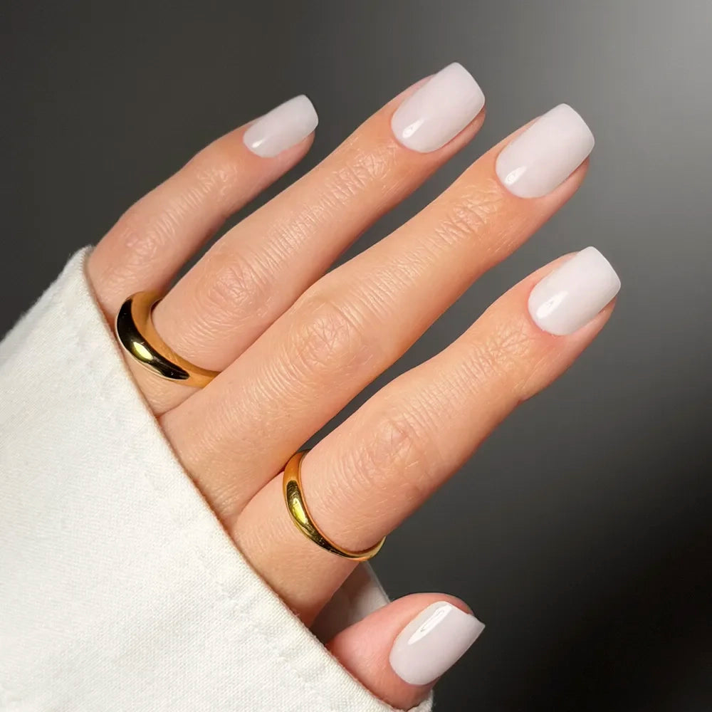 A hand with neatly manicured, short squoval nails in the shade Clean Cut by glamnetic wears two simple gold rings—one on the middle and one on the ring finger—against a neutral background, with a white shirt sleeve partially visible.