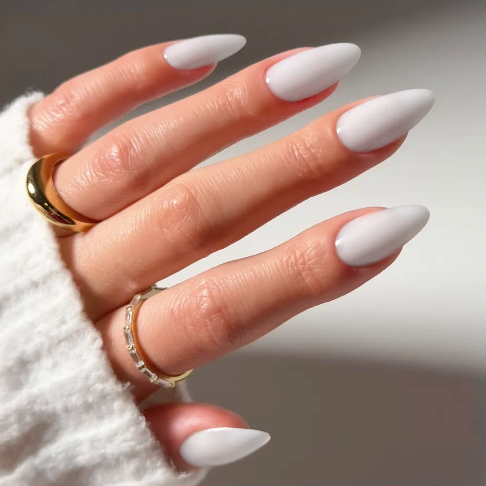 A hand with long, almond-shaped Clouded press-on nails by glamnetic, painted matte white, wears gold rings and a cozy white sweater against a soft background for a chic, salon-quality look.