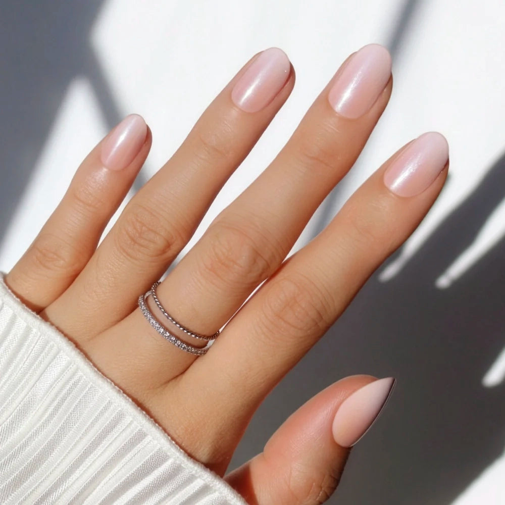 A hand with neatly manicured, short oval Crystal Glaze press-on nails by glamnetic in nude pink, wearing two thin sparkling rings on the ring finger. The person has on a white ribbed long-sleeve top, with light casting shadows in the background.
