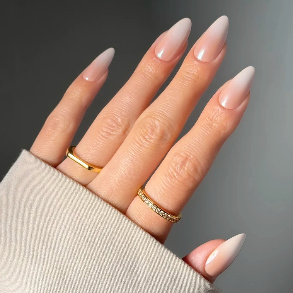 A hand wears glamnetic's Frosted Chai long, almond-shaped nude press-on nails with two gold rings—one plain, one with small stones—against a neutral background. The person is dressed in a beige sleeve. - Product image