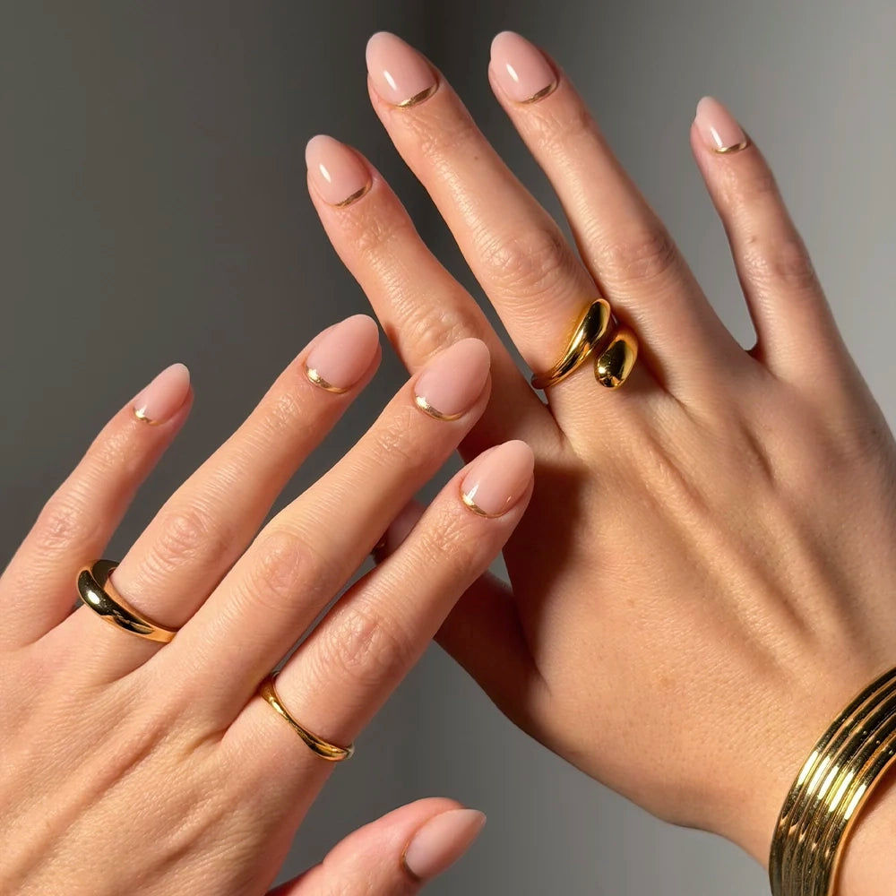 A close-up of two hands with short oval nails painted in glamnetic’s Glam Goals, a nude-pink shade with minimalist gold French tips. The manicured hands, accessorized with gold rings and bracelets, rest on a neutral background.