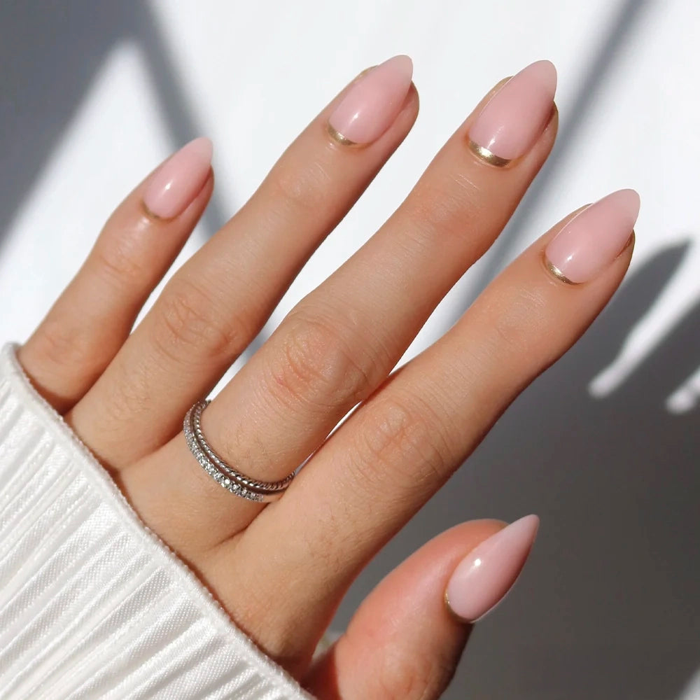 A hand models glamnetic's Goal Chaser press-ons: short almond nails in pale pink with thin gold French tips, paired with a ribbed white sweater and two stacked silver rings accented with small diamonds.