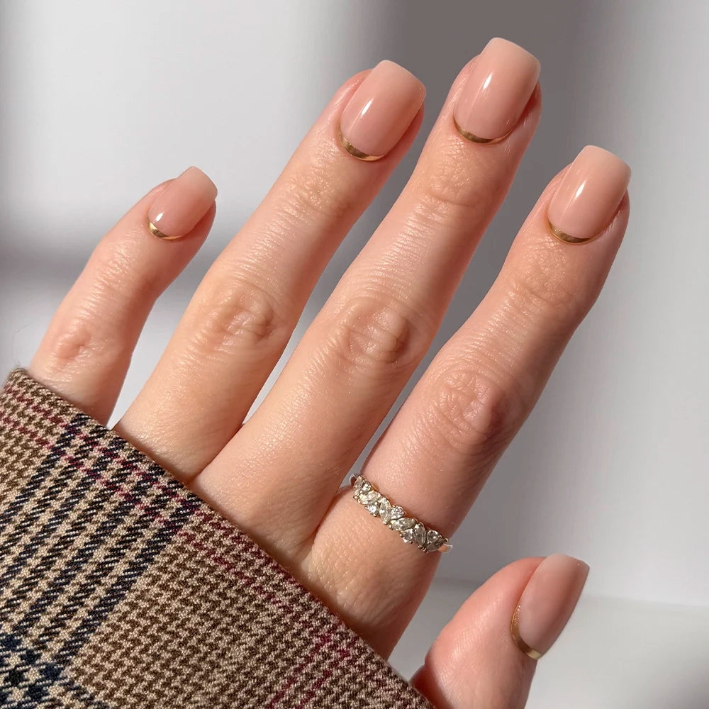 A hand with short squoval "Goal Setter" nude-pink gold French tip nails by glamnetic, wearing a plaid sleeve and a silver ring with small diamonds on the ring finger.