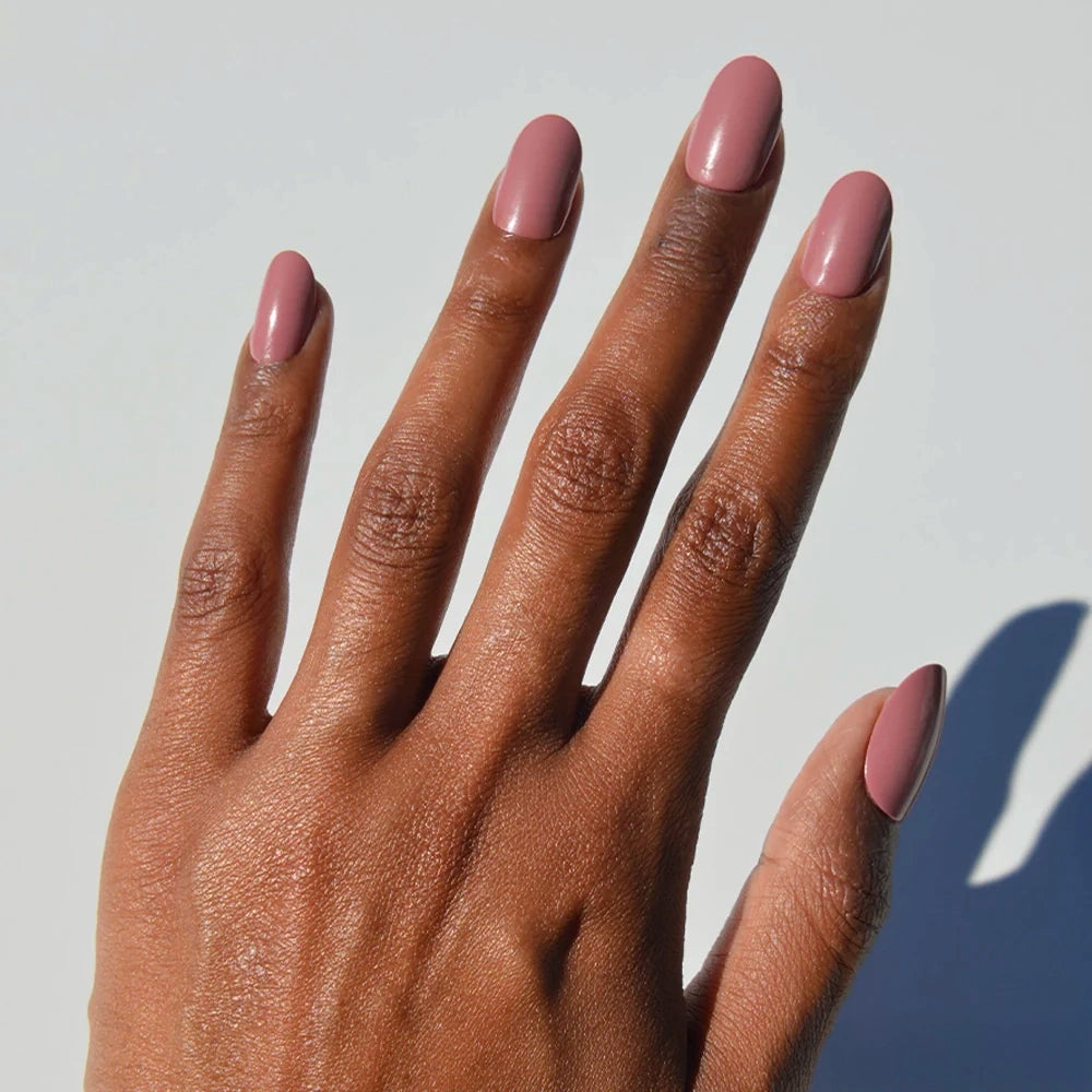 A hand with medium brown skin and short, neatly manicured nails painted in glamnetic’s “Mauve Mood,” a glossy muted pink-mauve shade, is shown against a plain light background, highlighting the salon-quality manicure.