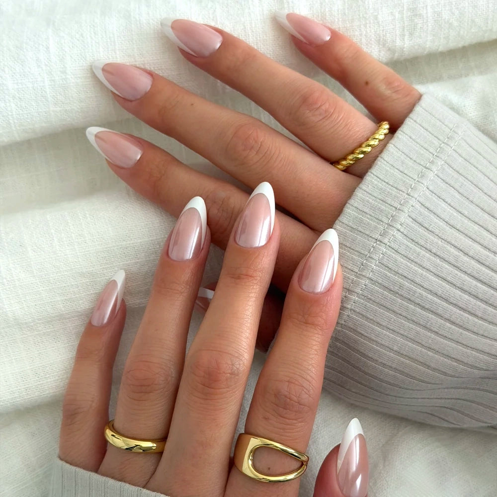 Hands with long, almond-shaped French tip nails in glamnetic’s Mother Pearl, featuring a glossy glazed finish. The person wears gold rings, and a light-colored ribbed sleeve can be seen in the background. - Product image