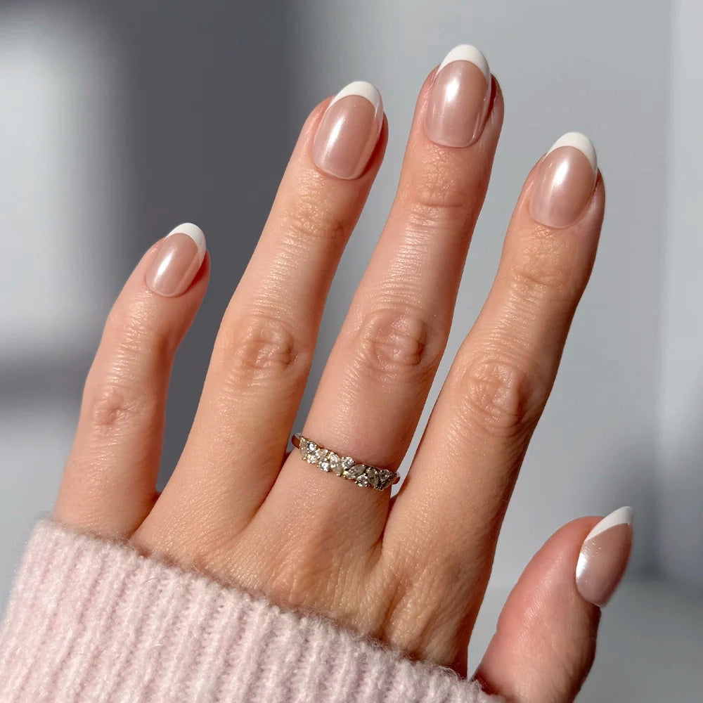 A hand with a Pearlfect French tip manicure by glamnetic shows off a silver ring with multiple small diamonds on the ring finger, set against a soft neutral background. The person wears a light pink ribbed sweater. - Product image