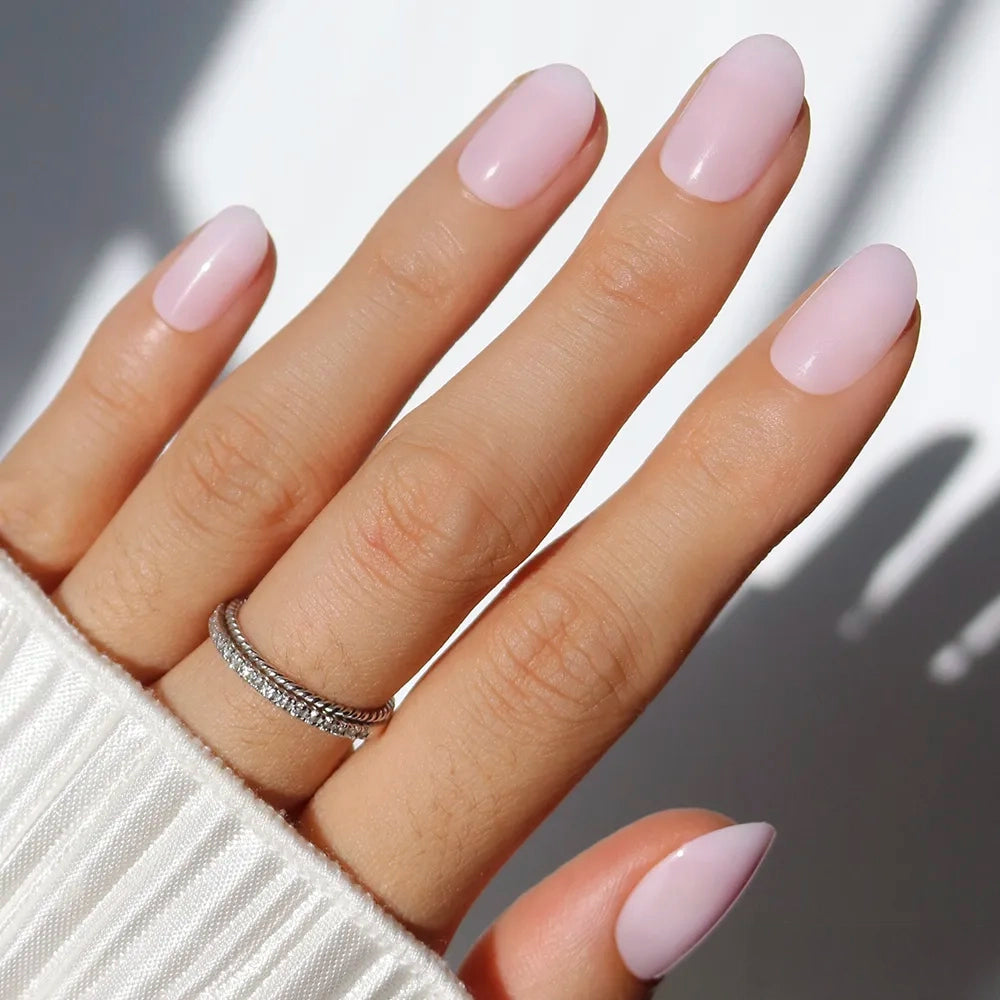 A hand with oval-shaped nails painted in Pink Blush by glamnetic showcases a salon-quality mani. A silver ring with small stones adorns the finger, while a white ribbed sleeve partially covers the wrist.