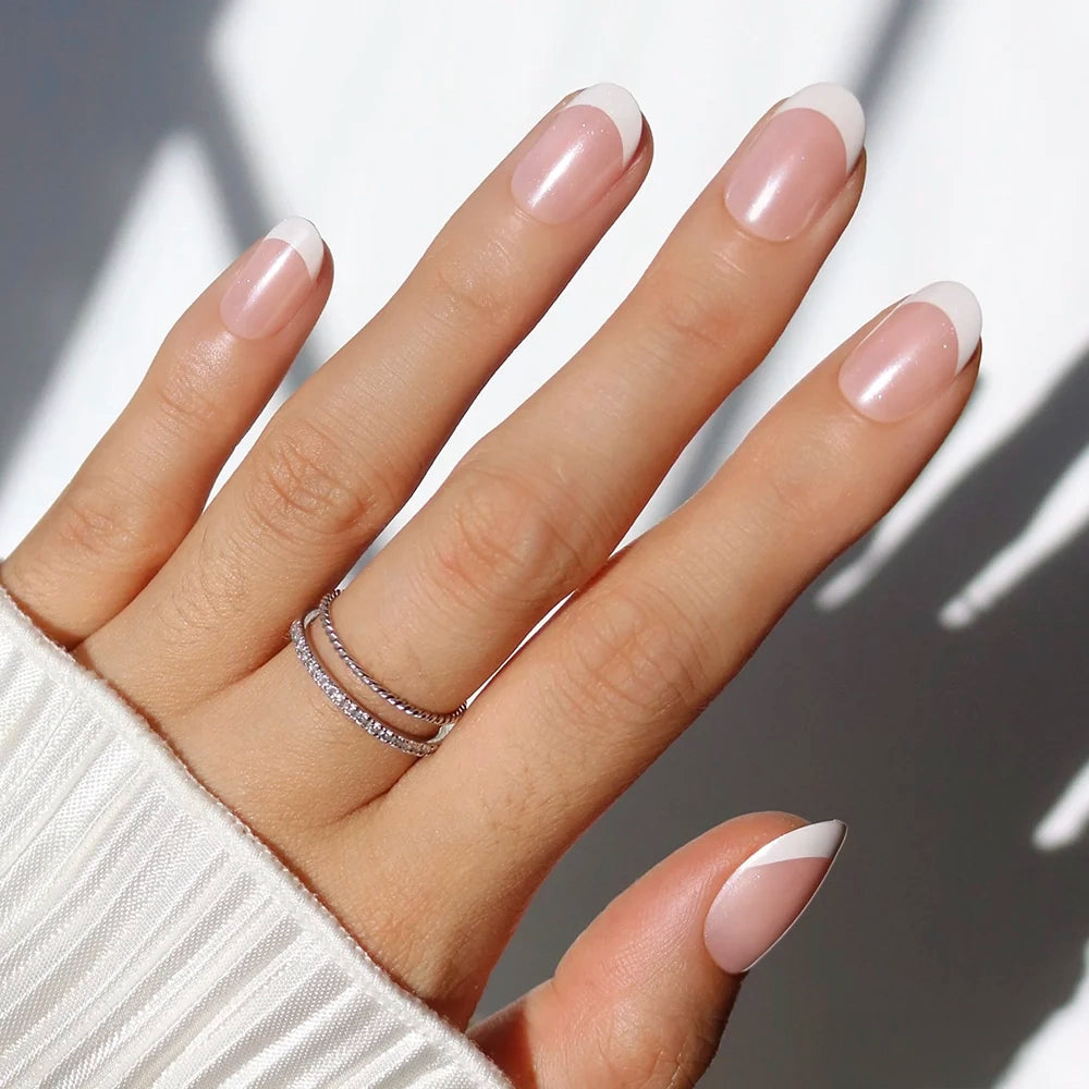 A hand with a light skin tone wears glamnetic's She's a Pearl press-on nails in a minimalist French tip. The almond-shaped, pale pink nails have white tips, are teamed with silver rings on the ring finger, and a white ribbed sleeve.