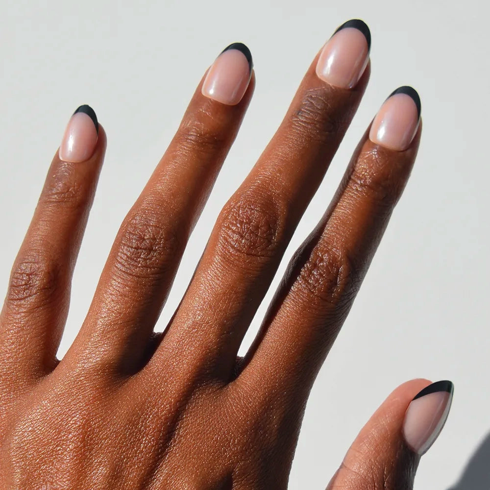 A close-up of a hand with medium brown skin wearing glamnetic Smoke Salt press-on nails featuring a nude base and thin black French tips, displayed against a plain white background.