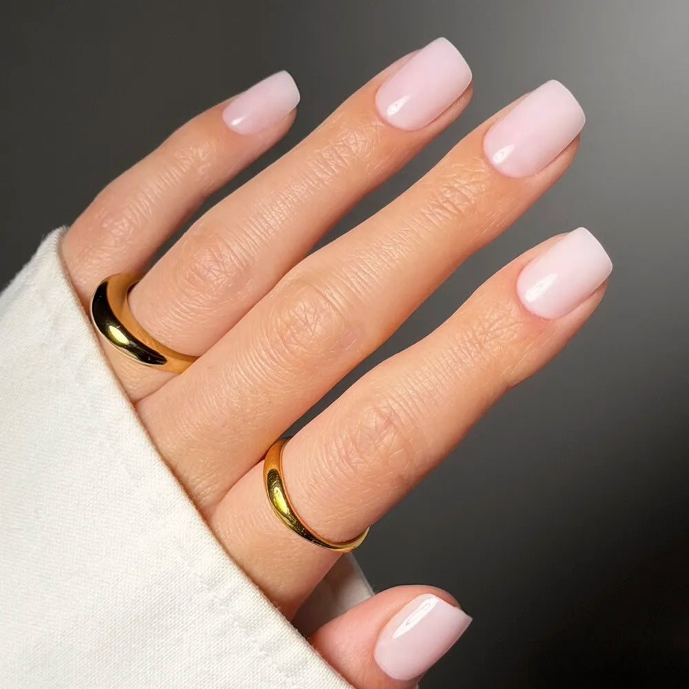 A hand with neatly manicured Soft Girl press-on nails by glamnetic, in light pink short squoval style, wears two shiny gold rings on the index and ring fingers. The nails peek from a white sleeve against a dark, blurred background.