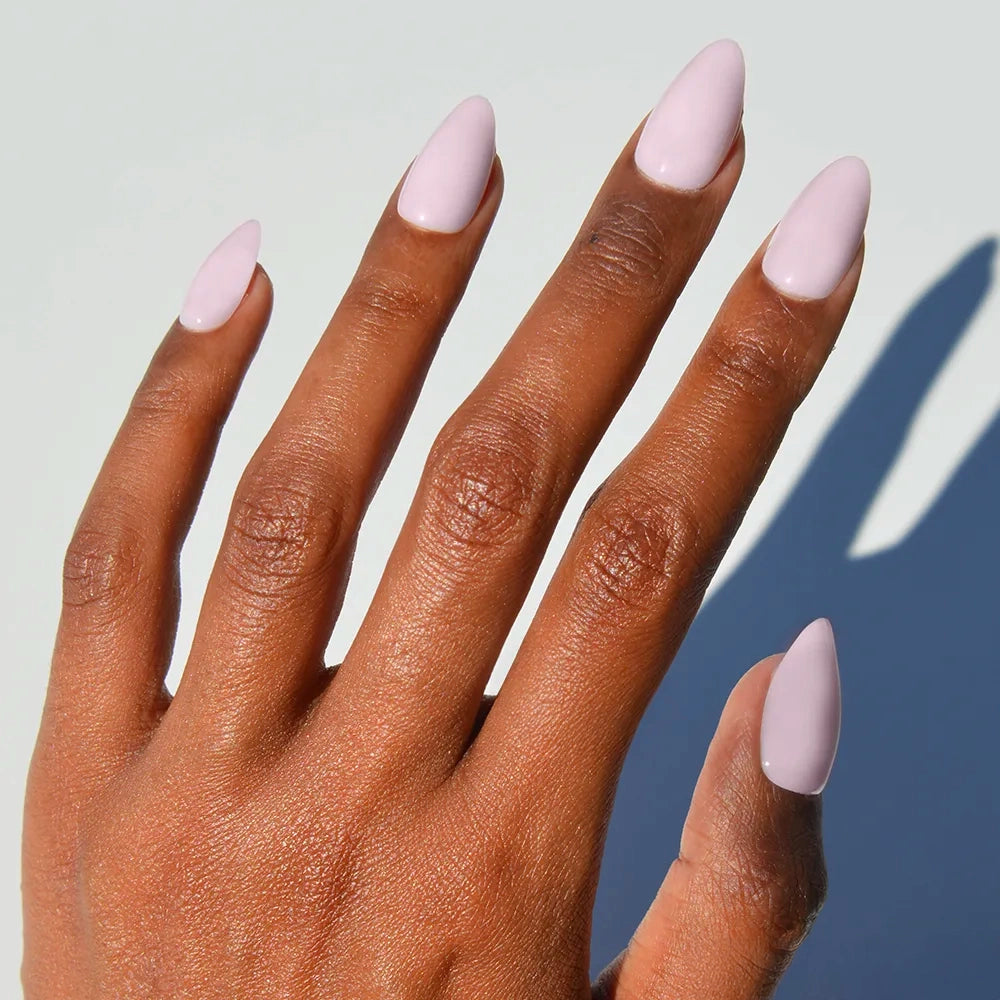 A hand with medium brown skin wears glamnetic's Sugar Doll press-on nails, featuring long almond shapes in pale pink, set against a simple light background.