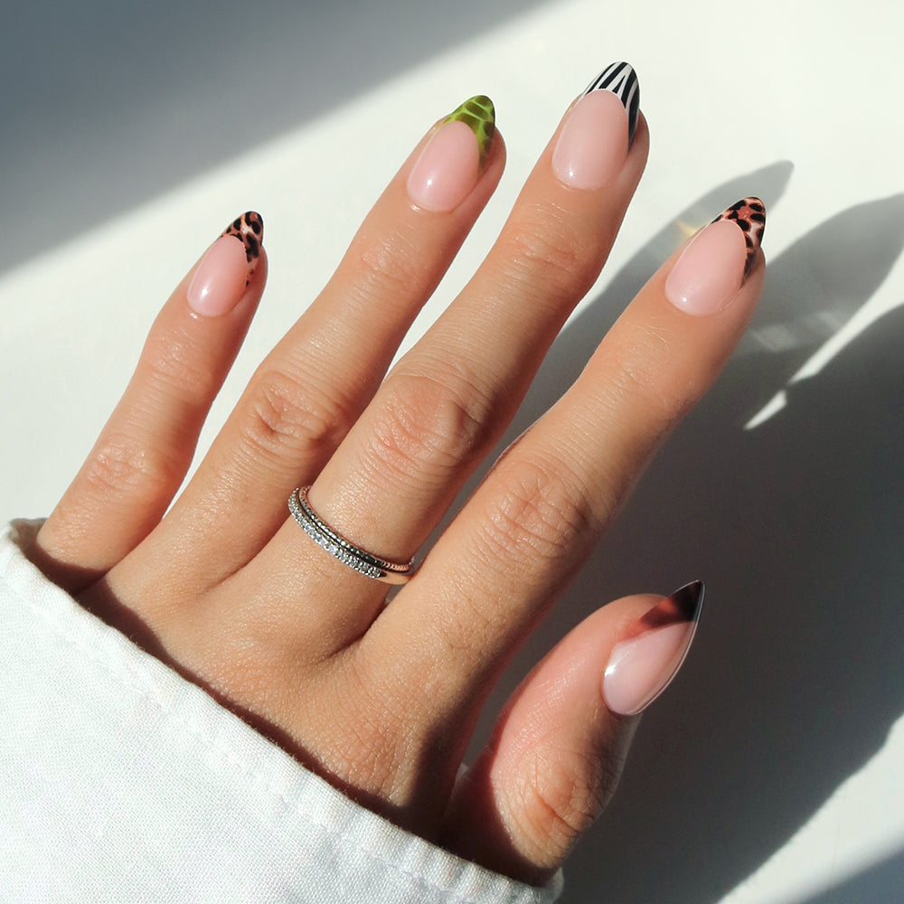 A hand with long almond-shaped 4 THE GLAMSQUAD nails by glamnetic features green leaf and black-and-white stripe nail art on a nude base. The person wears two stacked rings, with a white sleeve partly covering the wrist.
