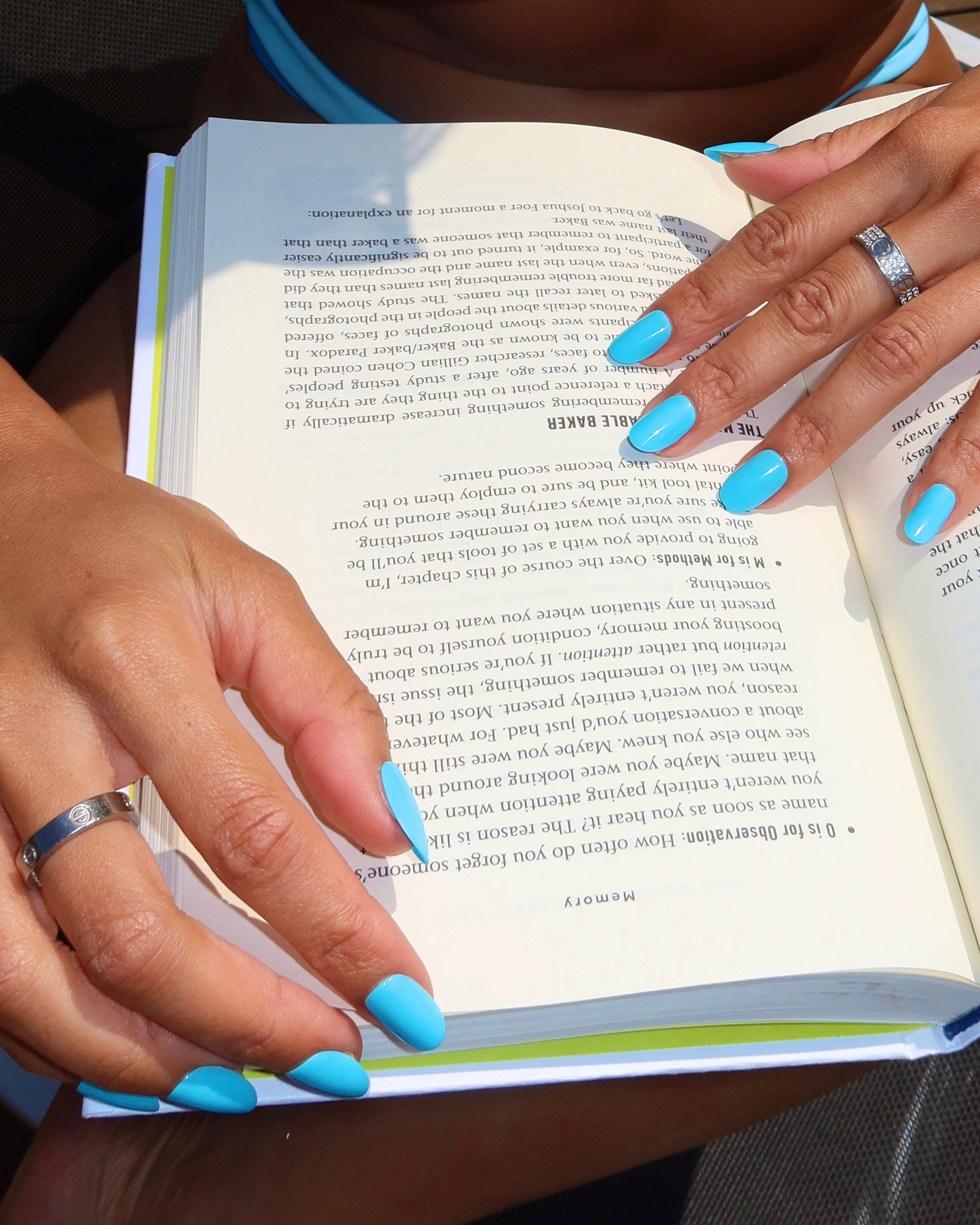 A person with long, glamnetic Azure Blue nails and silver rings holds open a book while reading outside in the sunlight. The text appears upside down to the viewer, capturing a vibrant moment of summer fun. - Product image