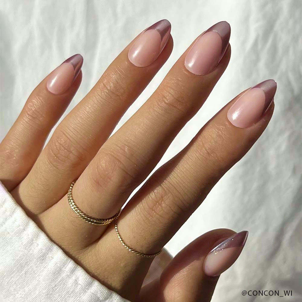 A hand with short almond-shaped nails showcases glamnetics Rock Candy Nail Set—a minimalist French manicure featuring a soft pink base and chrome rose gold tips. Two delicate gold rings and a white long-sleeve shirt complete the elegant look.