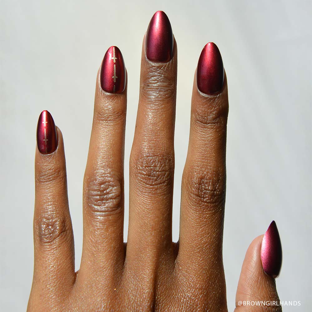 A hand with medium brown skin shows off a short almond manicure painted in metallic burgundy, accented with minimalist gold lines on the middle and ring nails, capturing the Gryffindor™ look by glamnetic.