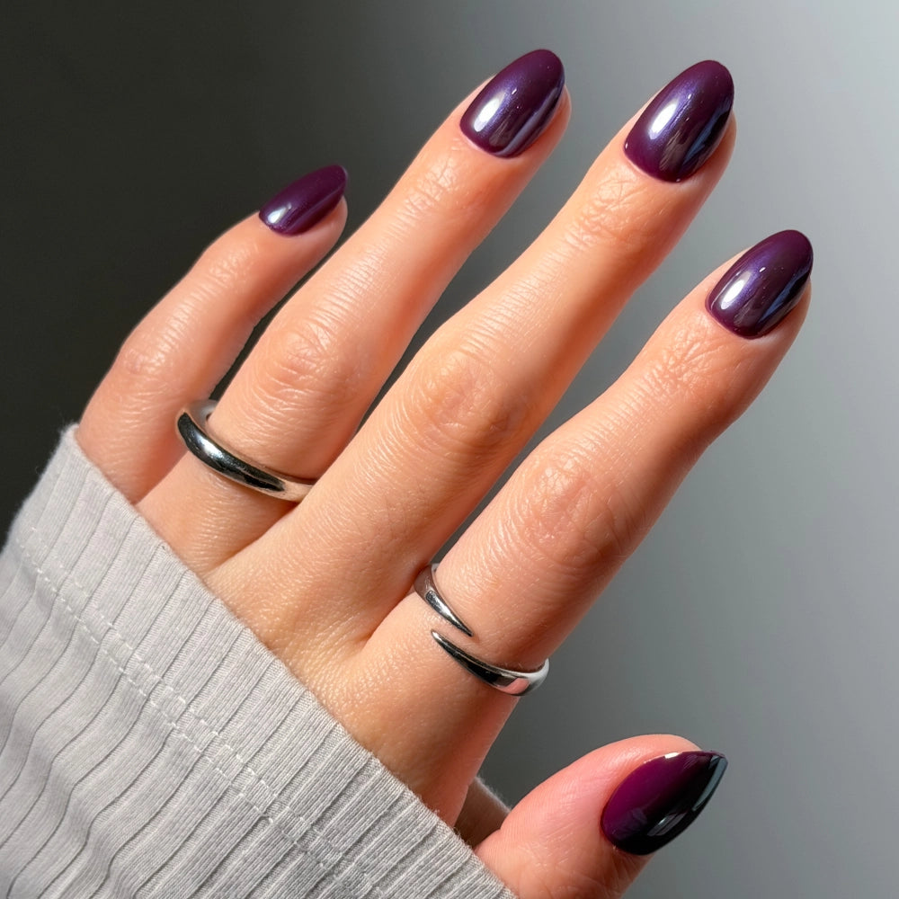 A hand showcases glamnetic’s “After Hours,” a shiny, dark purple nail polish with a sleek, glazed finish on short oval nails. Three silver rings adorn the fingers, and the person wears a long-sleeve, light gray ribbed shirt against a soft gray background. - Product image