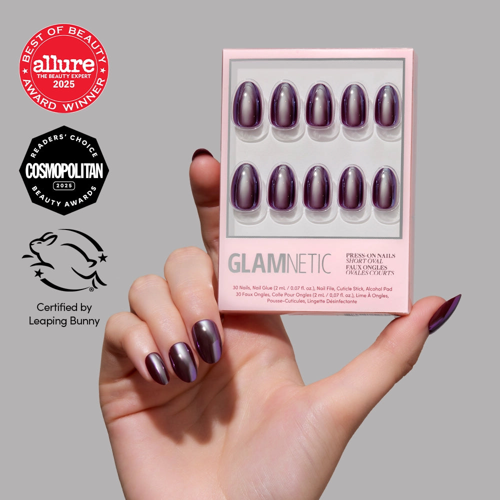 A hand with shiny, purple short oval press-on nails holds a glamnetic After Hours nail kit. The kit displays ten matching nails with a glossy, glazed finish. Beauty award badges from Allure, Cosmopolitan, and Leaping Bunny are visible on the left side. - Product image