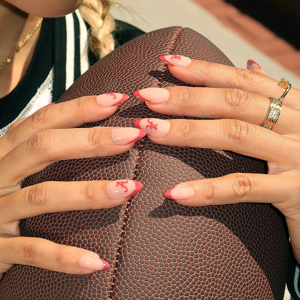 A person with long, almond-shaped glamnetic Alabama Crimson Tide Press-On Nails—pink with red tips and small red letters—holds a textured brown football. They wear a ring, black-and-white jacket, and have a blonde braid. - Product image