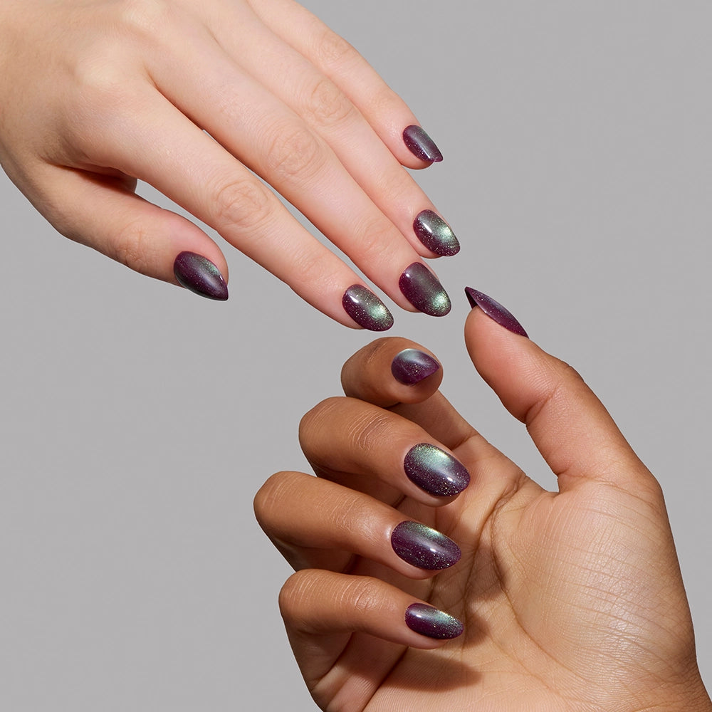 Two hands with short oval nails, painted in glamnetic’s “Alakazam”—a shimmery purple and green gradient—reach towards each other against a plain gray background. The velvet finish of Alakazam catches the light beautifully, adding an extra touch of elegance to the scene. - Product image