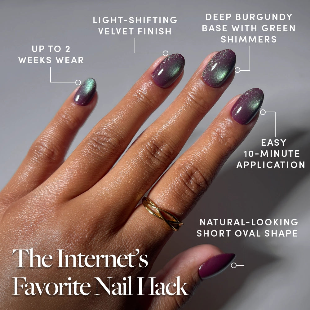 A hand showcases short, oval burgundy “Alakazam” nails by glamnetic, adorned with a green shimmer and a rich velvet finish. The text highlights their light-shifting effect, up to 2 weeks of wear, quick 10-minute application, and natural appearance. A gold ring graces the middle finger. - Product image