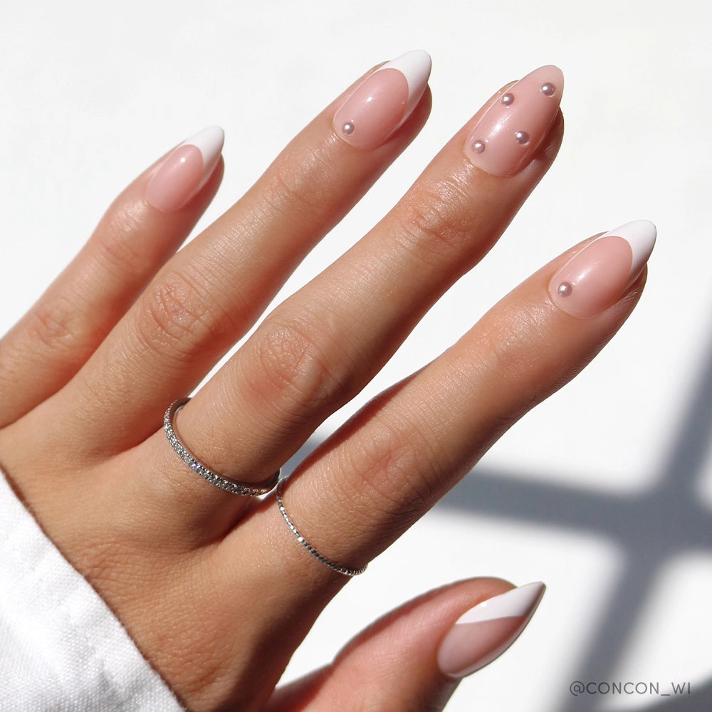A hand displays almond-shaped nude pink press-on nails with a classic white French tip design. Select nails are adorned with dainty pearl accents. The hand is accessorized with thin silver rings, and lit with soft natural sunlight.


